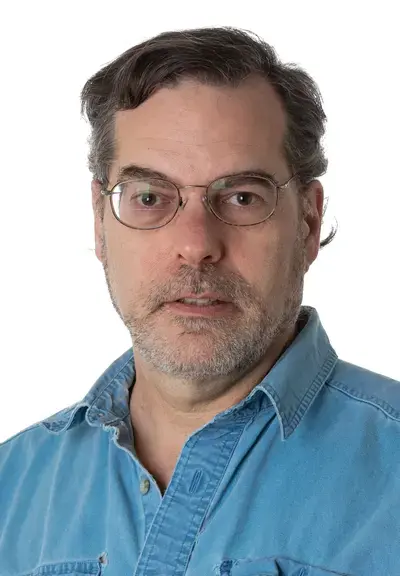 a white man with a denim shirt and glasses looking at the camera