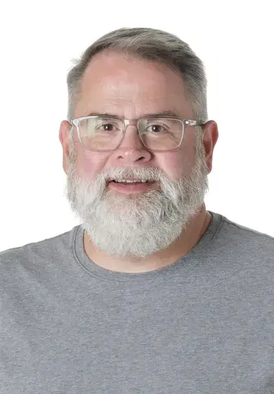 a white man with short white hair and a white beard smiling at the camera, he is wearing glasses and a grey shirt