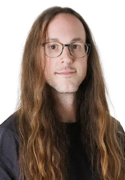 a white person with glasses and long brown wavy hair looking at the camera, they are wearing a black shirt
