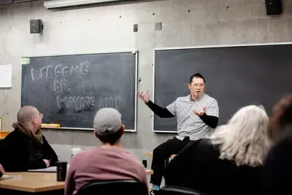 A man wearing a white dotted shirt sits casually in front of two blackboards, lecturing to classroom of students whose backs are to the camera. A chalk message on the blackboard says "Welcome Dr. Wayne Au!"