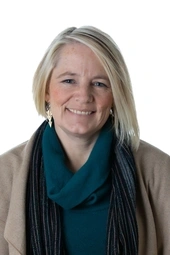 a white woman with blond bob smiling at the camera, she wears a brown cardigan and a teal turtleneck