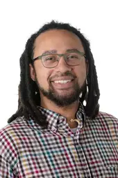 a black man with shoulder length dreads and a short beard smiling at the camera, he is wearing glasses and a multi-colored checkered shirt