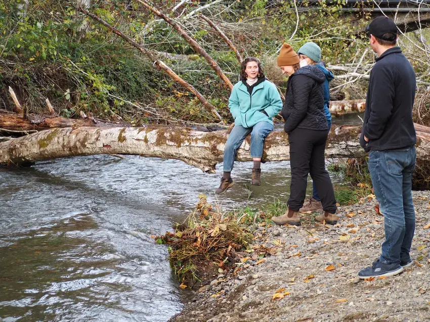 students outside next to a stream, one is sitting on a log facing the camera, the others are facing each other speaking