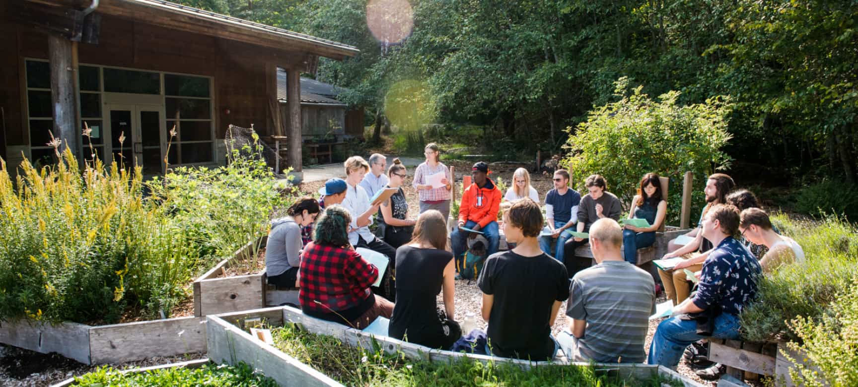 Photograph of students of all ages and races, holding a discussion on chairs in a circle outside amongst shrubs and bushes. A large wooden cabin is in the background.