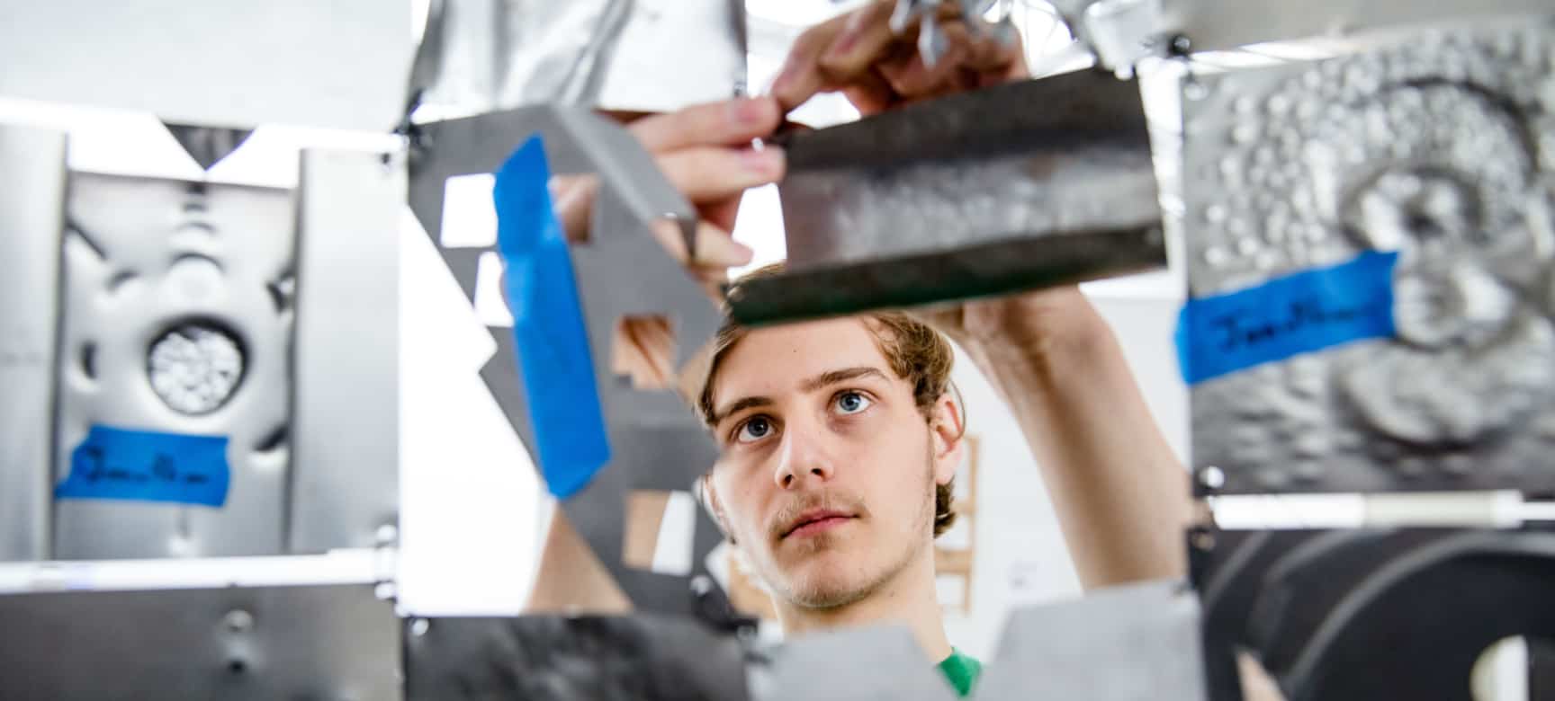 A male student is featured in focus as he holds up a piece of metal for examination. Other metal components are shown as blurred in the foreground.